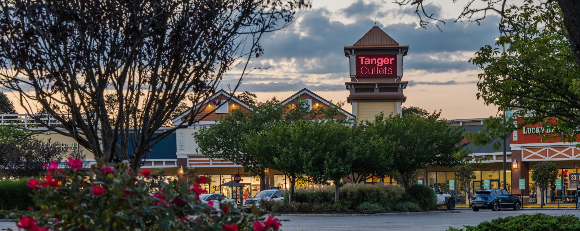 Outlet mall entrance with a "Tanger Outlets" sign, surrounded by trees and shrubs. Cars parked in front, under a cloudy evening sky.