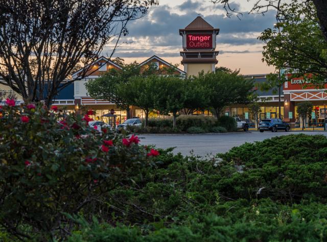 Outlet mall entrance with a "Tanger Outlets" sign, surrounded by trees and shrubs. Cars parked in front, under a cloudy evening sky.