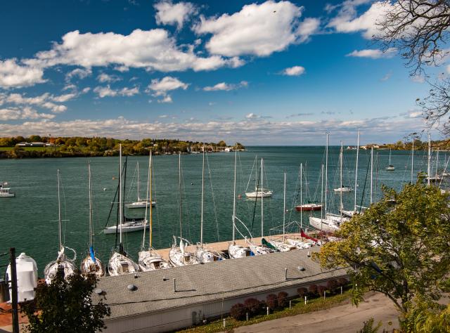 A marina with numerous sailboats docked along the waterfront, overlooking the Niagara River under a partly cloudy sky. Trees and a road are visible in the foreground.