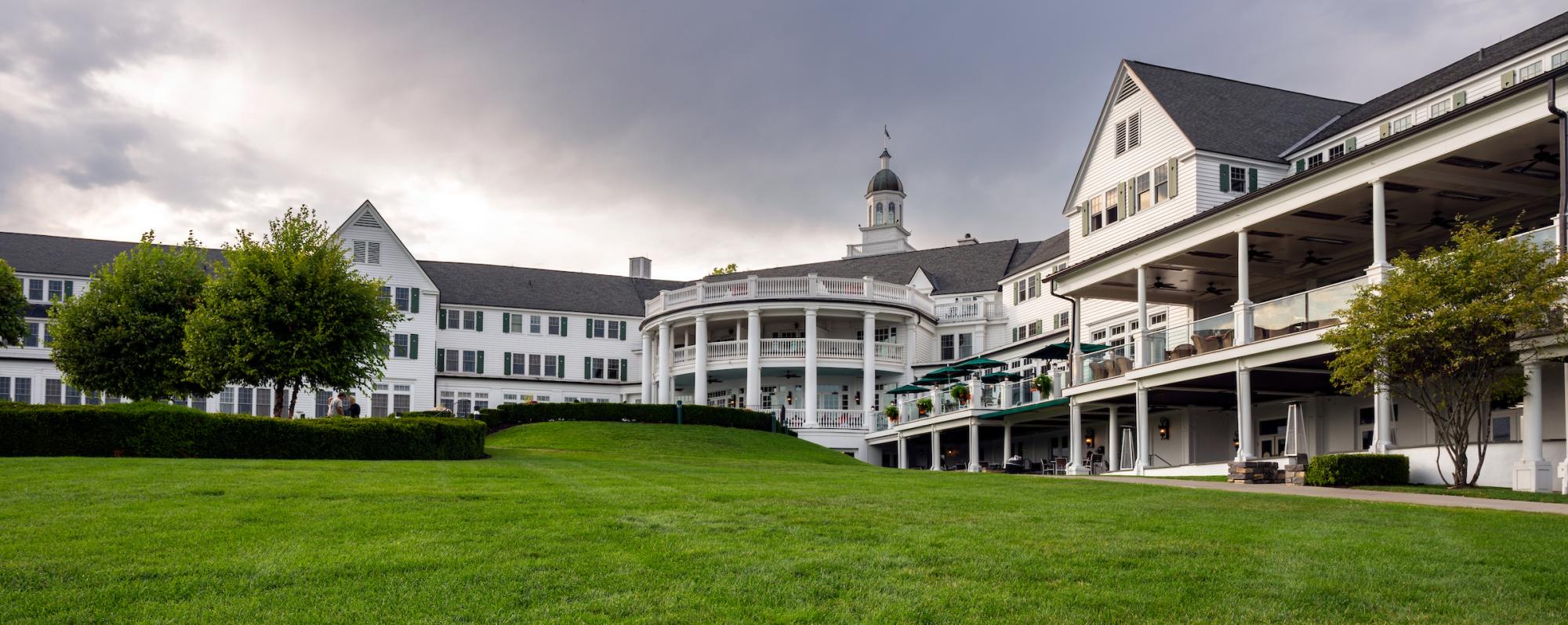 A lush green lawn in front of the grand Sagamore Resort, a white building with multiple stories and a circular porch, under a cloudy gray sky.