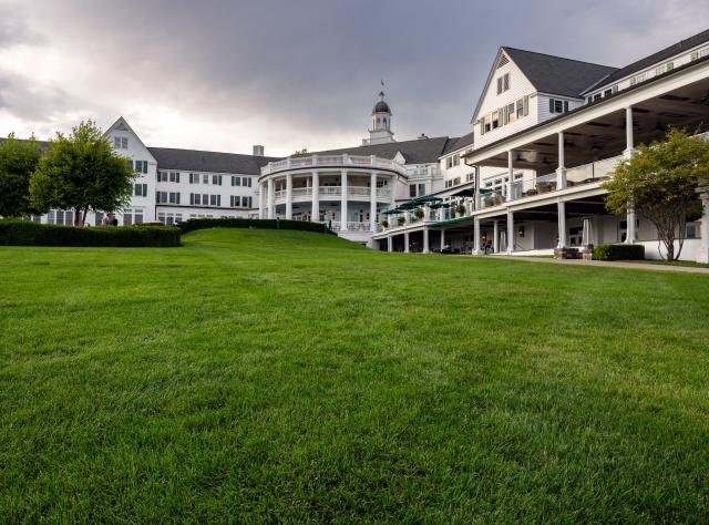 A lush green lawn in front of the grand Sagamore Resort, a white building with multiple stories and a circular porch, under a cloudy gray sky.
